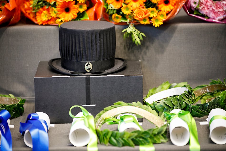 En svart hatt som står på en låda, omgiven av blommor och hoprullade diplom. Foto.