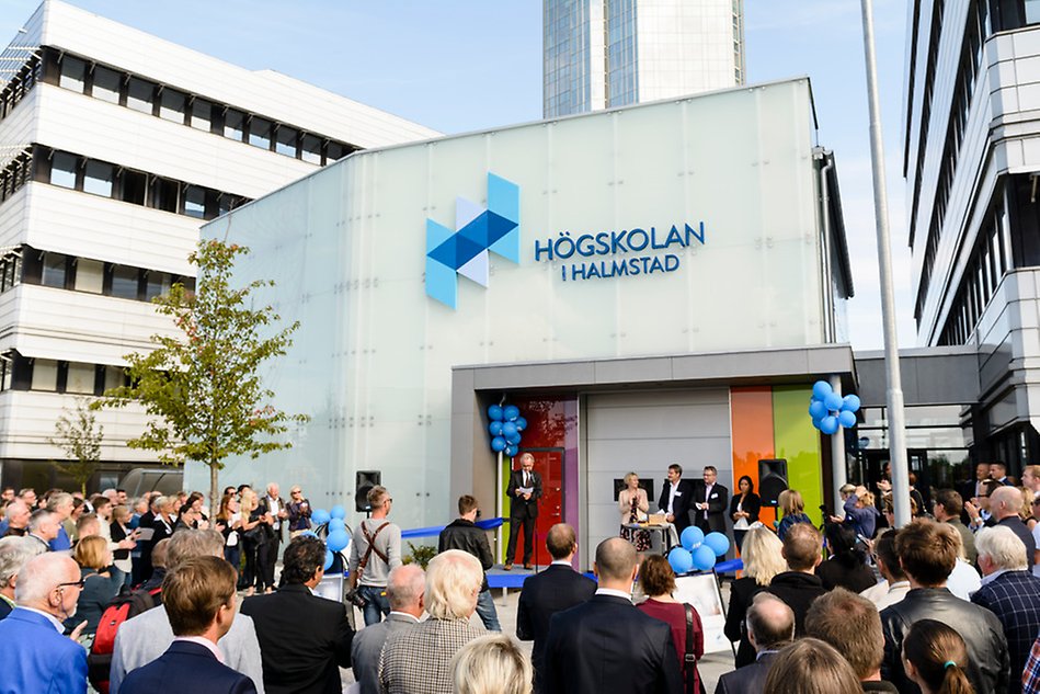 Stort antal åhörare framför en vit kvadratisk byggnad med Högskolan i Halmstads logotyp. Foto.
