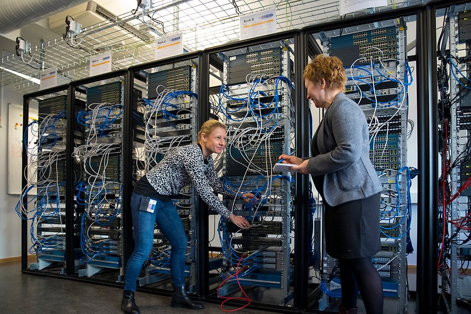 Two women stand in front of a server rack and show cords and computers. Photo. 