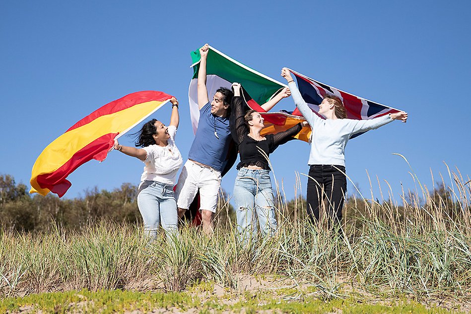 Tre unga kvinnor och en ung man står bredvid varandra på en sanddyn. De ler och har flaggor i sina händer som de viftar med. Foto.