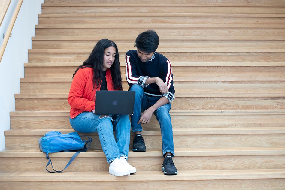 Two people sitting on a large staircase, looking at one laptop. The people look relatively young and one of them has a backpack. Photo. 