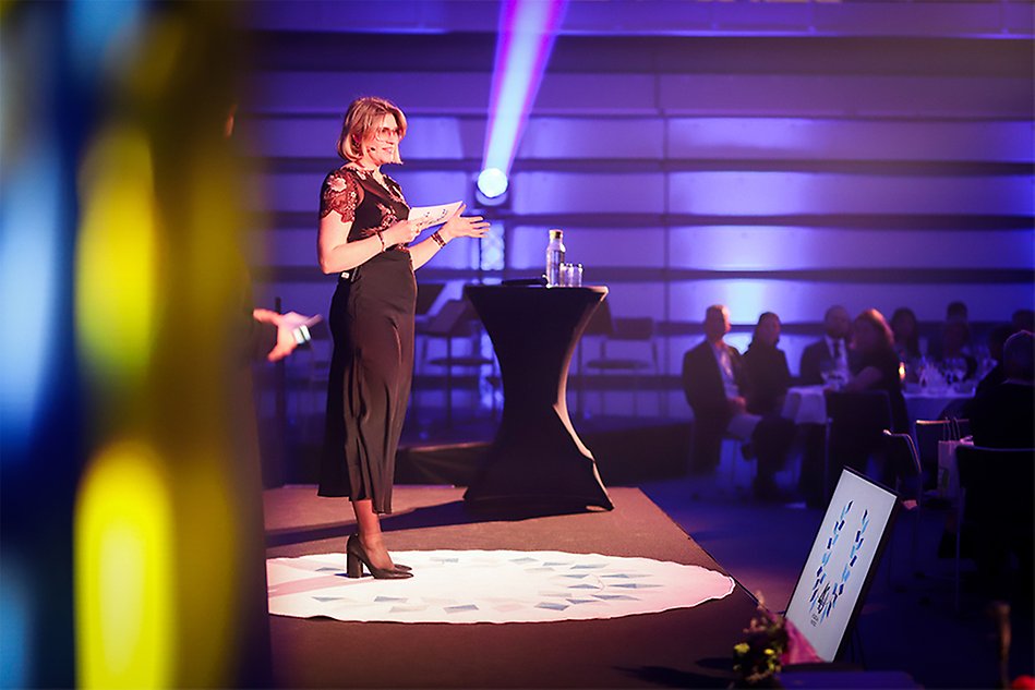 En kvinna med ljust hår och glasögon och en mörk klänning står på en scen. I sina händer har hon ett talarkort. Foto.