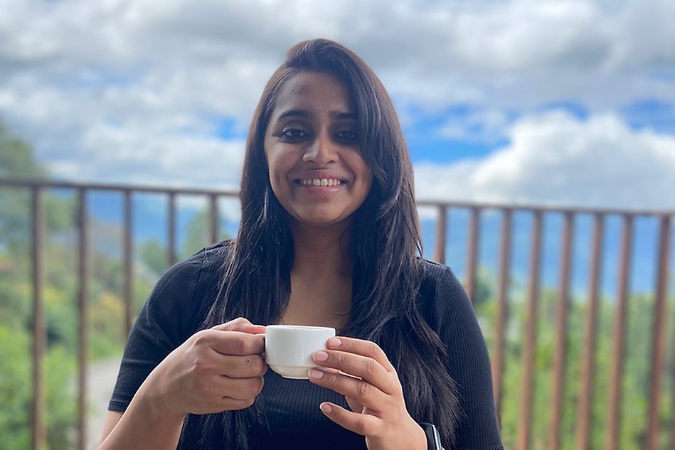 Person with long dark hair holding coffee cup, smiling. Blue sky behind. Photo.