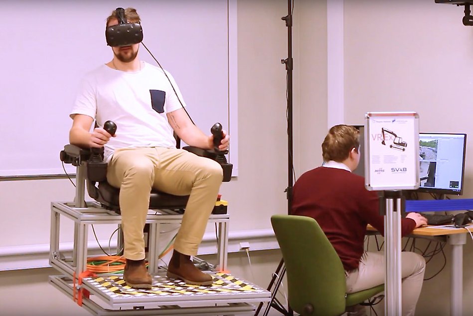 En man sitter i en fåtölj med VR-glasögon och en styrspak i vardera hand, en annan sitter vid en dator.