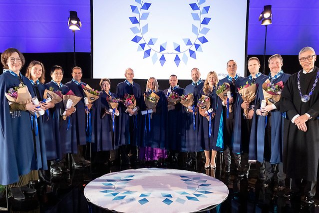 En stor grupp människor i caper och med blommor händerna står i halvcirkel runt en rund matta. I bakgrunden en bildskärm med en blå dekoration. Foto