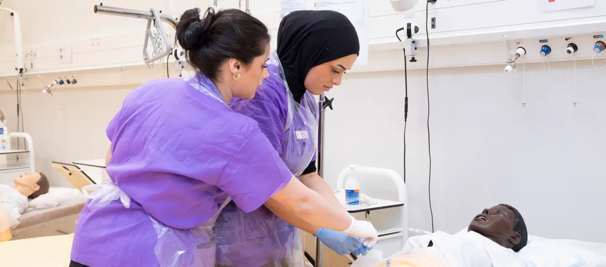 Två kvinnliga sjuksköterskor tar ett prov på en docka som ligger i en säng. Foto.