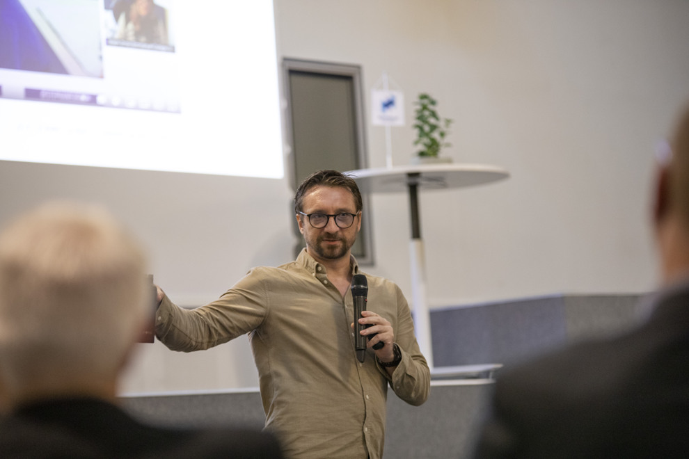 Man står med mikrofon och håller i en miniföreläsning inför ett antal människor. Foto.