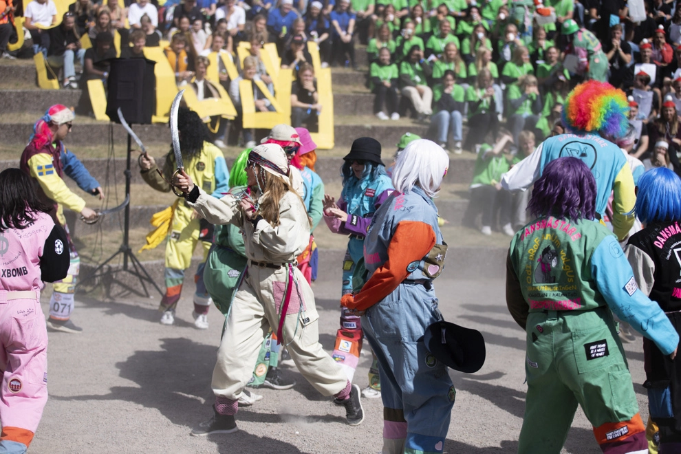Ett flertal människor i masker och färgglada overaller dansar inne på en amfiteater utomhus. Foto.