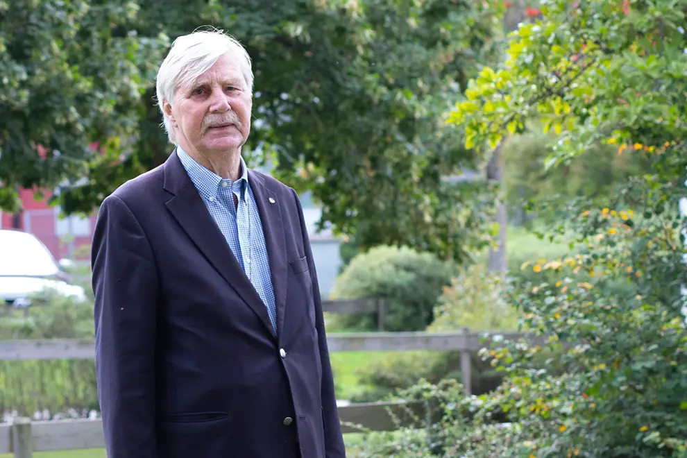 Person står vid träd och grönska utomhus och tittar in i kameran.