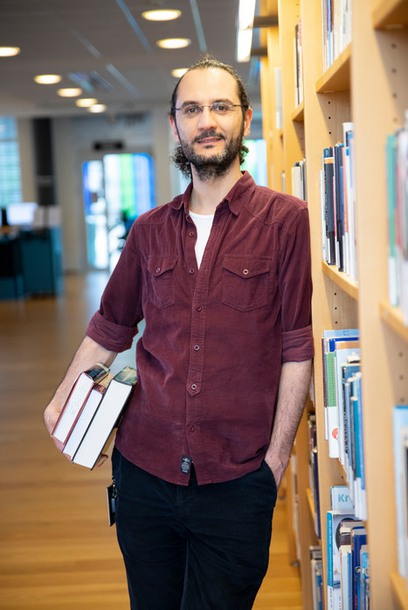 Man holding some books, leaning against a bookshelf. Photo. 