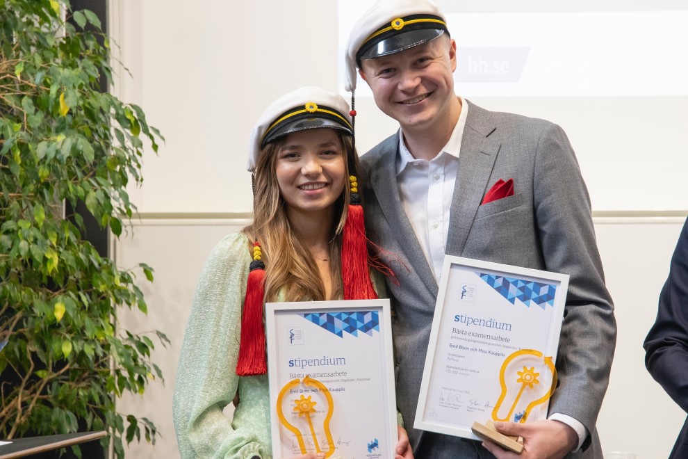 Två studenter ler mot kameran och håller i sina diplom och statyetter.