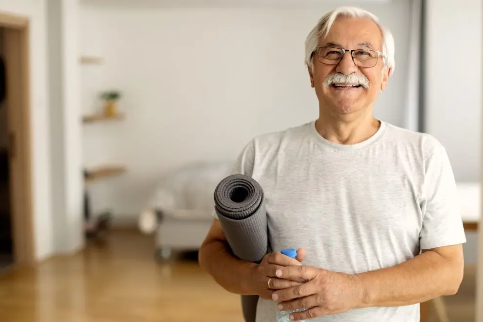 En äldre person med en yogamatta och en vattenflaska i handen. Foto