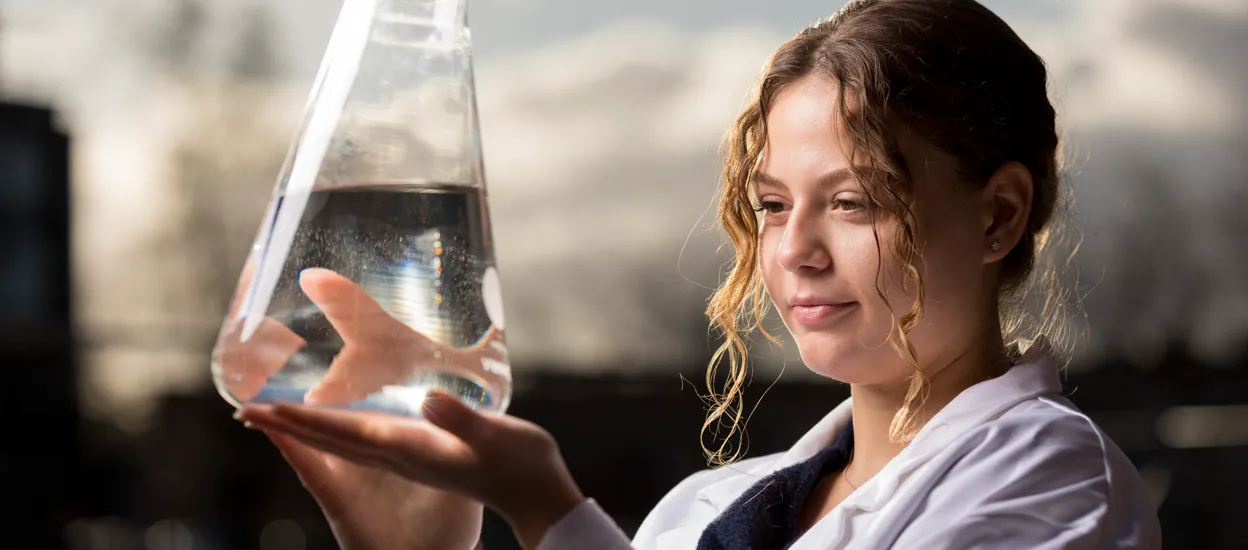 A woman dressed in a lab coat holds up an e-flask filled with clear liquid. Photo.