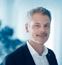 Man with short gray hair and beard and light button up looking into the camera.