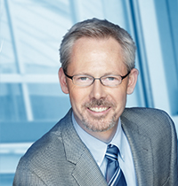 Man with grey hair and small glasses wearing grey suit smiling into the camera.