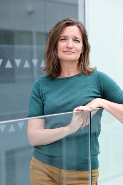 A woman wearing a green top standing in front of a brick wall with her arms folded. Photo.