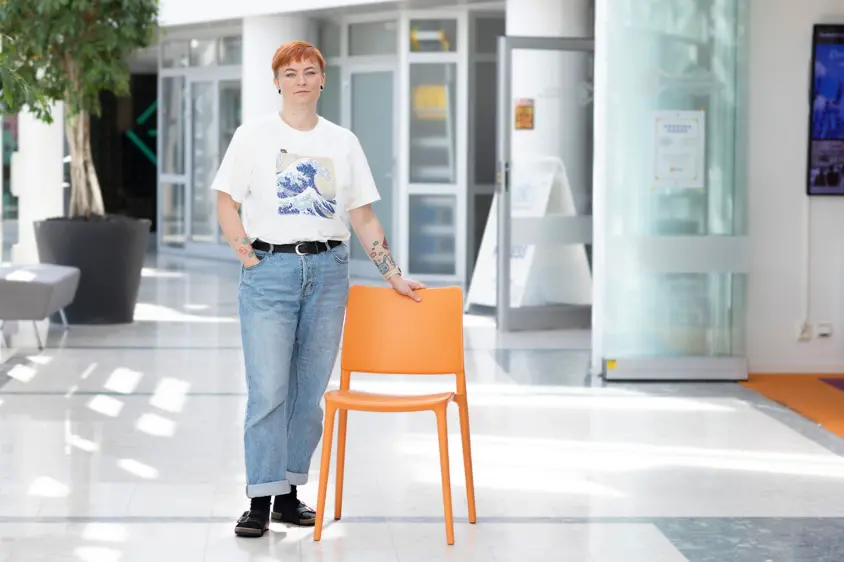 Person i jeans och t-shirt står i en ljus korridor. Personen har handen på ryggstödet på en orange stol. Foto.