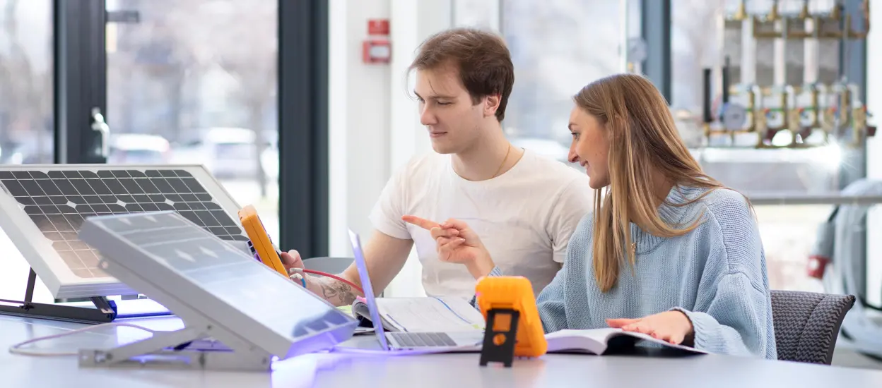 Två unga studenter en kille och en tjej sitterr vi ett bord med solcellspaneler och tittar på en dator. Foto.
