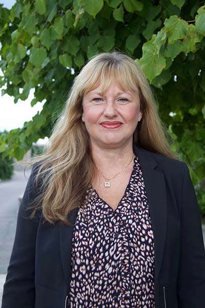 A woman standing looking into the camera and smiling. Greenery in the background. Photo.