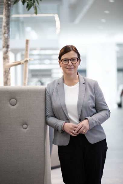 Woman in a grey jacket standing in a bright space, leaning on the back of a piece of furniture. Photo.