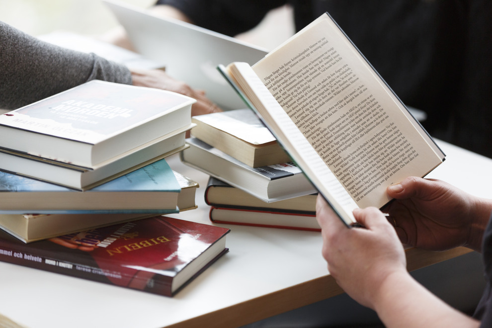 Böcker som ligger staplade. En bok är uppslagen som två händer håller i. Foto