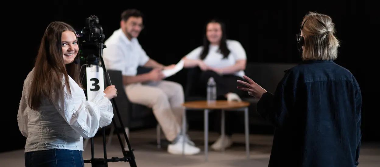 En ung kvinna står i en inspelningsstudio med medieutrustning och ler mot kameran.