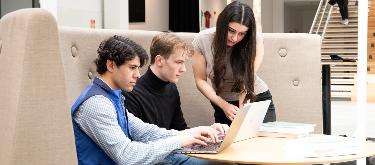 Three students gathered by a sofa at campus, working on their computers. Photo.