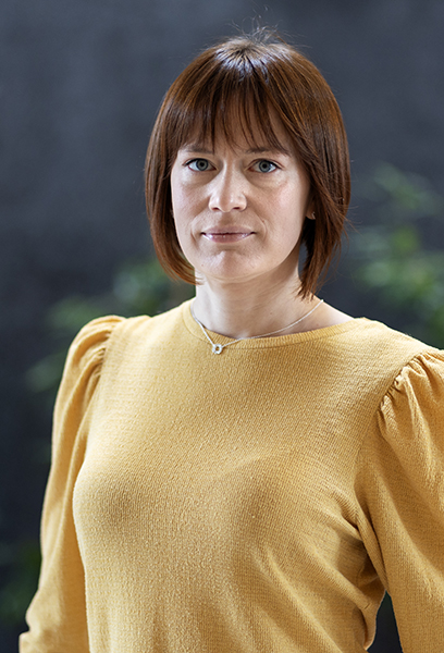 A woman in a yellow shirt looks into the camera. Blurry plats are seen in the dark background.