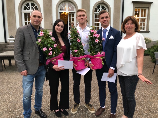 Fem personer ler och tittar in i kameran, tre av dem har blommor i famnen. Foto. 