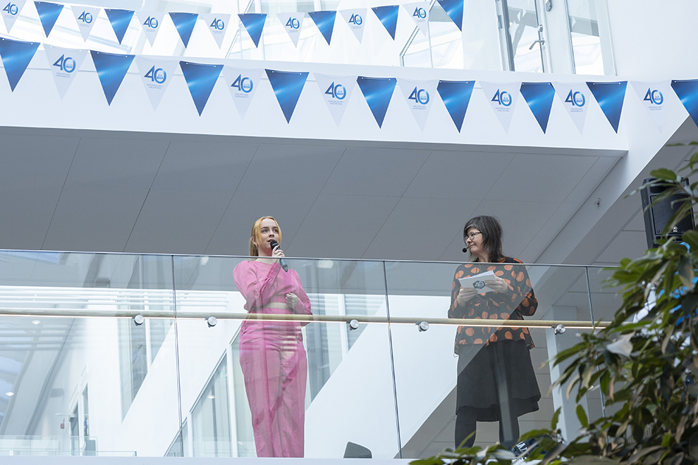 Woman in pink clothing talks in a microphone next to a woman on a balcony. Photo.