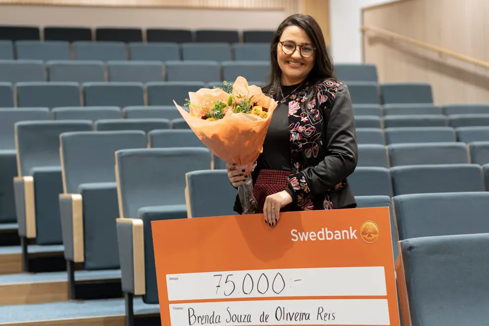 Kvinna i mörkt hår och glasögon står i en tom föreläsningssal. I ena handen har hon en blombukett och i den andra en stor värdecheck. Kvinnan ler mot kameran. Foto.