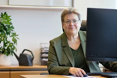 En person kikar fram bakom en datorskärm. I bakgrunden en växt. Foto