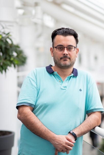 A man with dark hair wearing an aquamarine polo shirt is leaning against a railing. Photo.