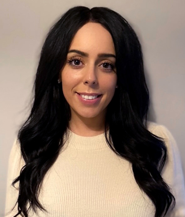 A woman with dark hair and light sweater looks into the camera. 