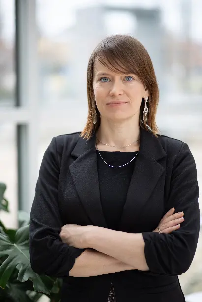 A woman stands in front of a window with crossed arms looking into the camera.