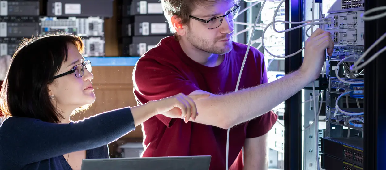 En kvinna sträcker sig och pekar mot några kablar och en man håller i en kontakt. Foto.