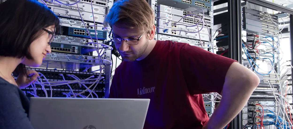 Två studenter står vid en skärm i en datorsal. bakom så finns det dator-rack med många sladdar. Foto.