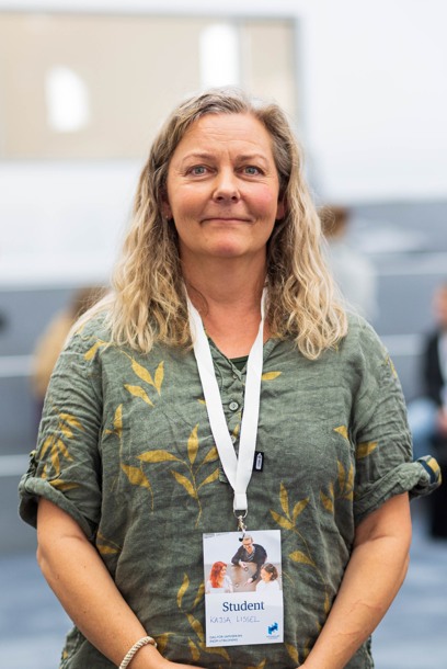 Kvinna med blont, lockigt hår och grön klänning med gula blad står i ett rum fyllt med studenter.