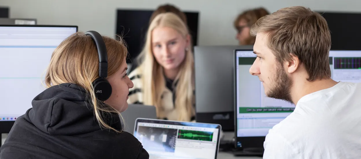 Två studenter sitter vid en varsin datorskärm och lutar sig mot varandra för att diskutera. En student i bakgrunden lyssnar på. De är i en datorsal. Foto.