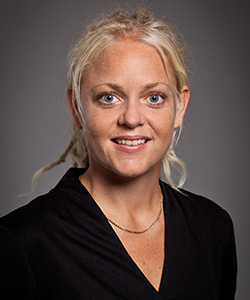 En kvinna med blont hår och mörk blus tittar in i kameran.