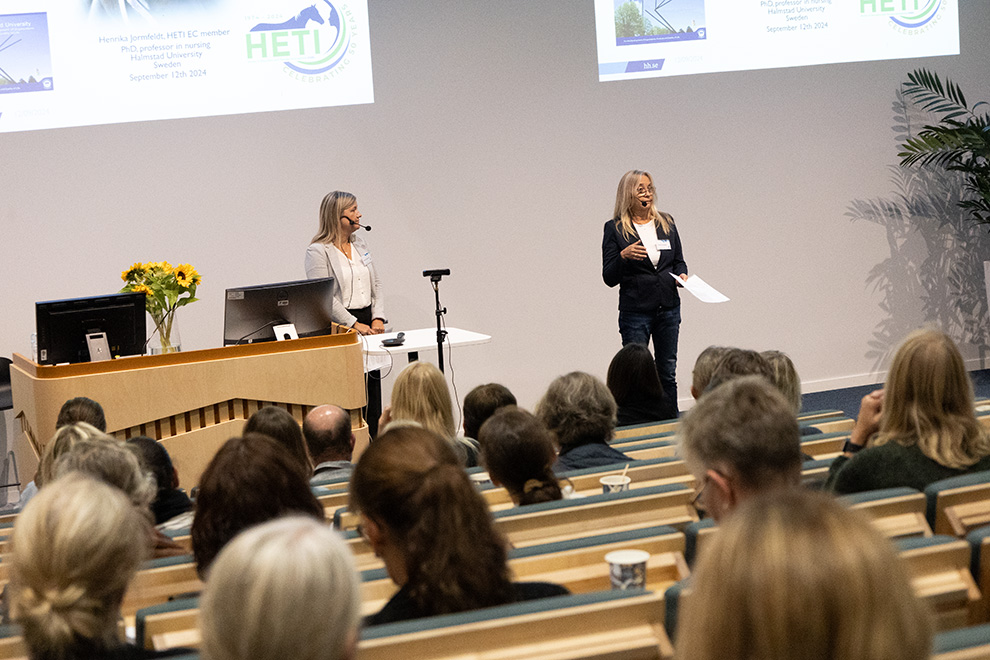 Två personer står framför en publik och pratar. Foto