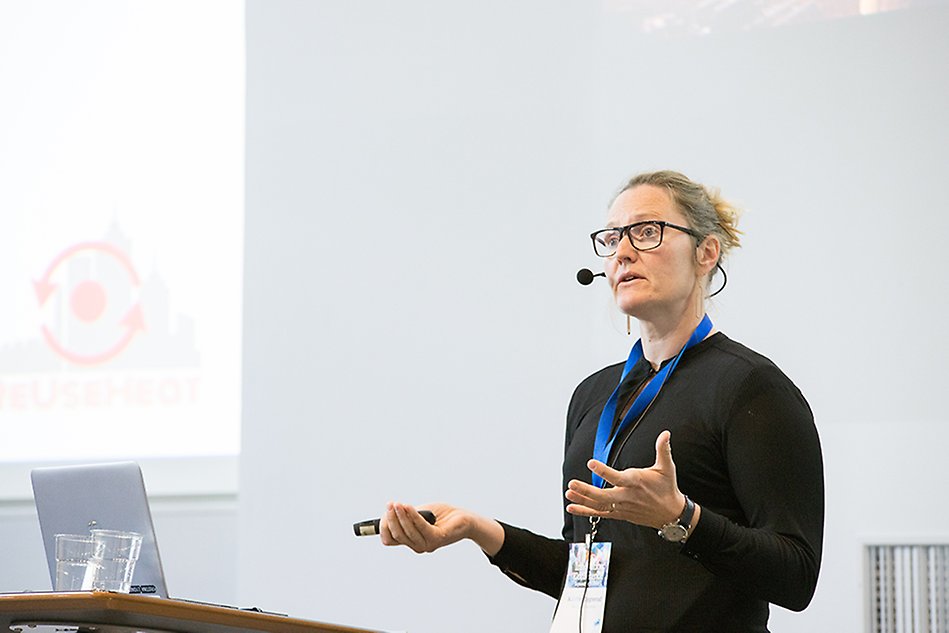 Woman standing to the right giving a speech. A screen with a drawn figure to left together with a computer and glasses. 