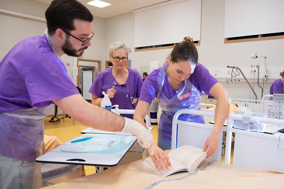 Sjuksköterskestudenter läser i en bok i en vårdrumsmiljö. Foto.