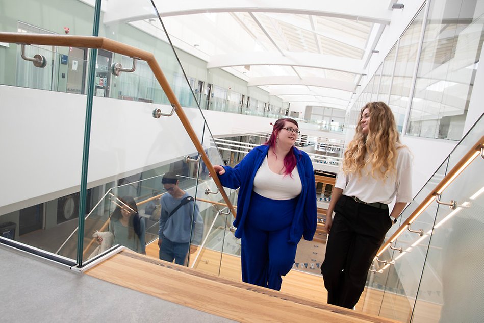 Two people are walking up the stairs in a bright building with glass windows. Photo.