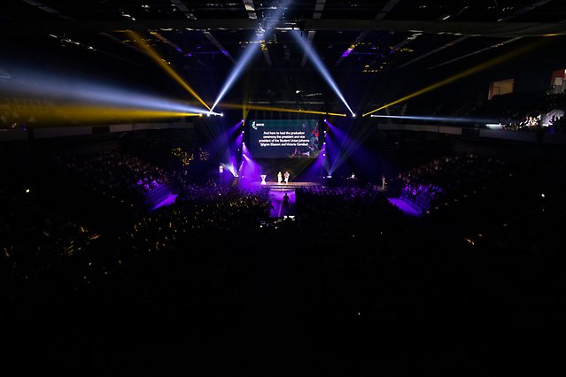 En fullsatt arena med strålkastare mot scenen. På scenen syns en stor monterskärm och två personer. Foto.