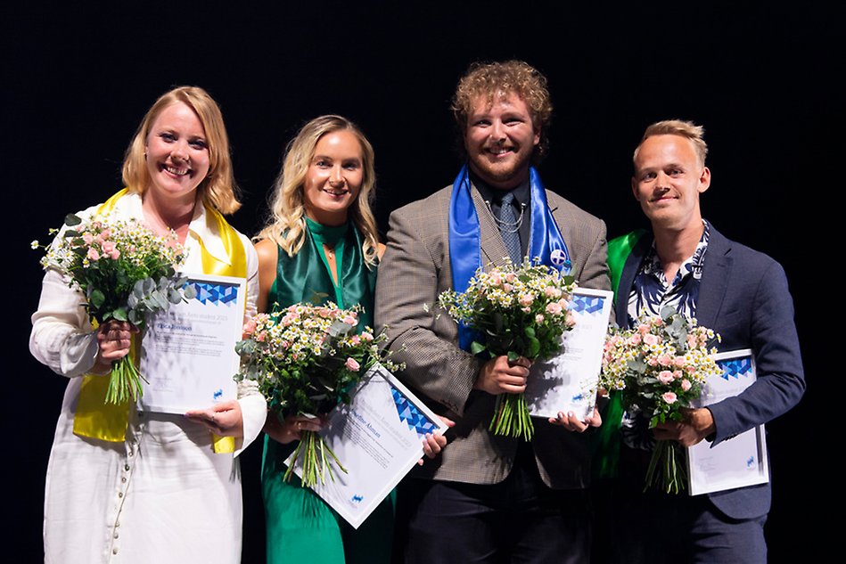 Two young women and two young men are standing in a row with flowers and diplomas in their hands. They are all dressed up. Photo.