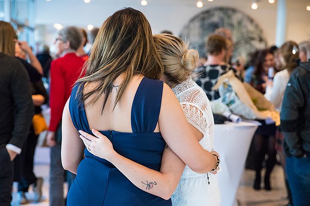 Two well-dressed young women are standing with their arms around each other’s backs with the backs against the camera. In the background there is a crowd of people. Photo.