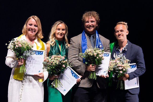 Two young women and two young men are standing in a row with flowers and diplomas in their hands. They are all dressed up. Photo.