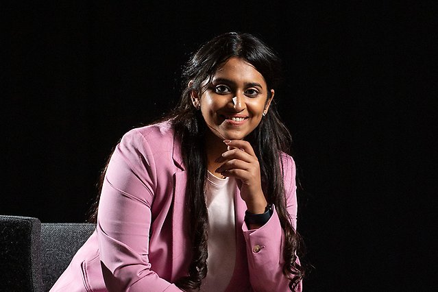 A woman in a pink jacket is sitting on a chair in a dark room. She is facing the camera smiling. Photo.