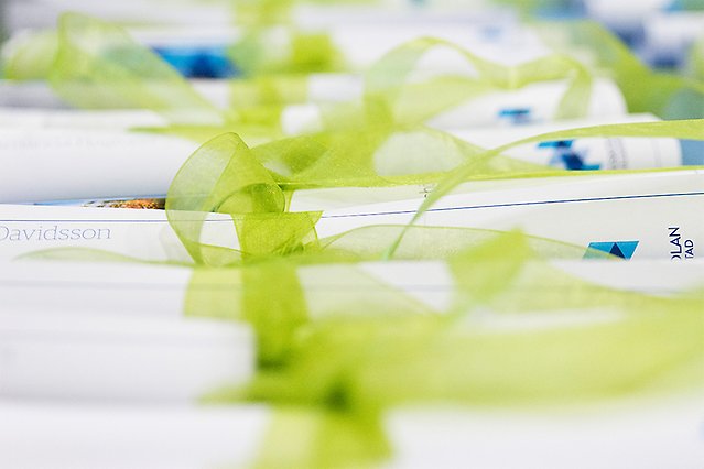 Several white and blue diplomas are lying rolled up on a table. Each diploma has a green ribbon tied around it. Photo.